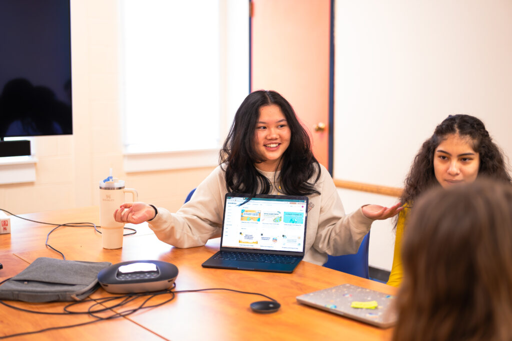 photo of woman sitting with her arms up showing the presentation on her computer, sitting at large table with other people next to her. a whiteboard and window and TV screen is visible in the background