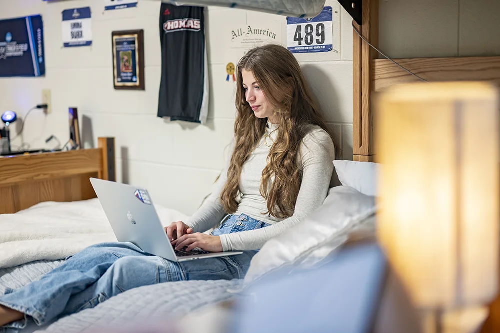 Thomas College student-athlete Emma Burr sitting in her dorm room working on a laptop at Thomas College in Waterville, Maine