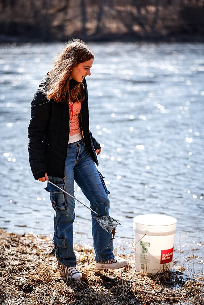 A student stands near a bucket with her instrument and the river in the background during Girls in Science Day 2026
