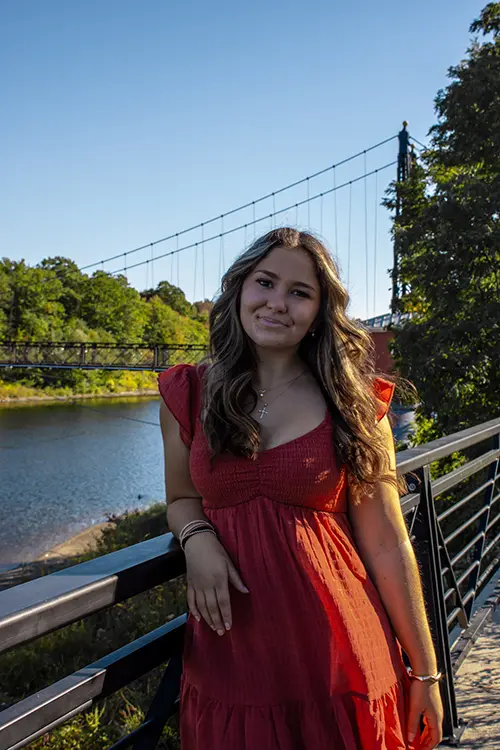 Thomas College student Alexis Spencer poses in front of a river in downtown Waterville with the walking bridge in the background