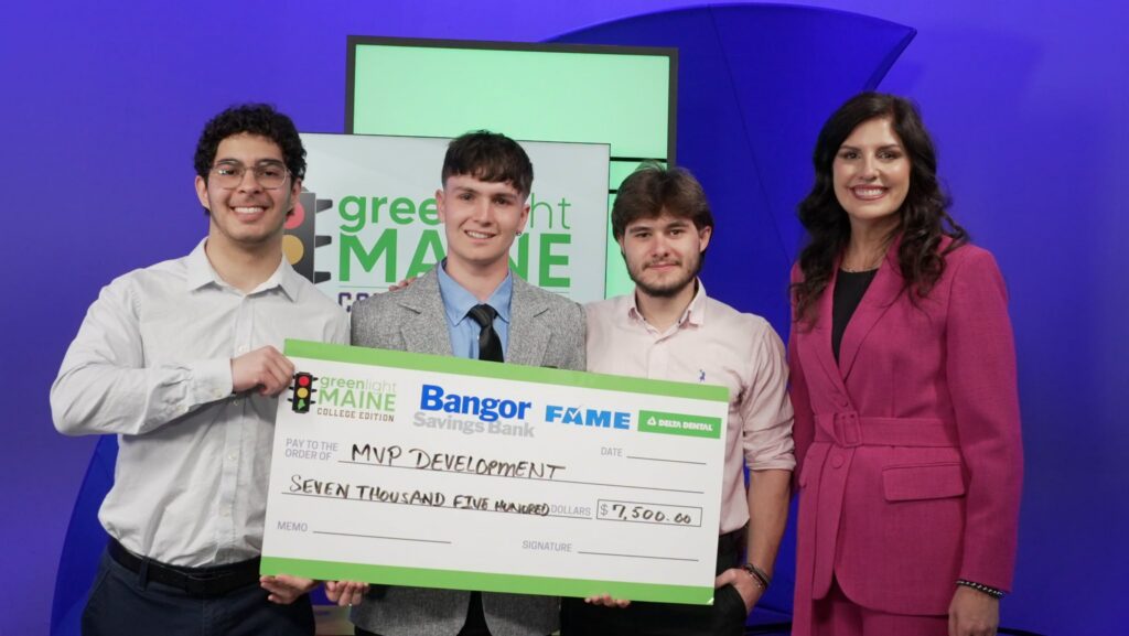Three Thomas College students stand with the host of Greenlight Maine holding a large check in a studio during the Greenlight Maine show