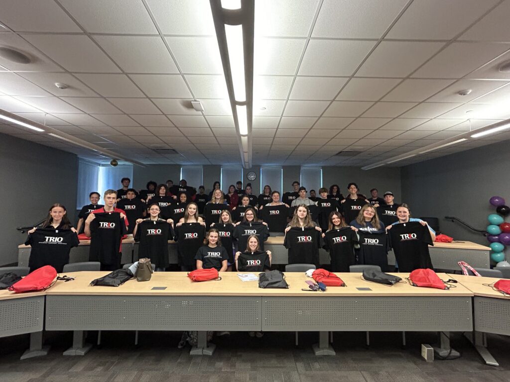 A group of students sit in a classroom holding their t-shirts up that read TRIO