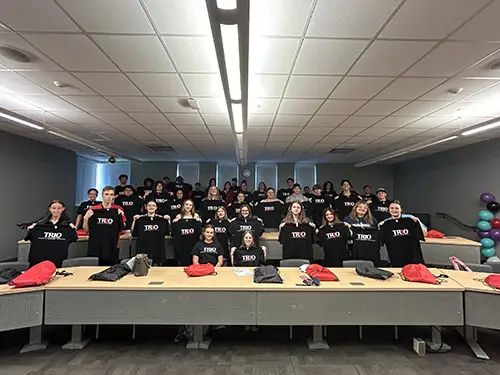 A group of students sit in a classroom holding their t-shirts up that read TRIO