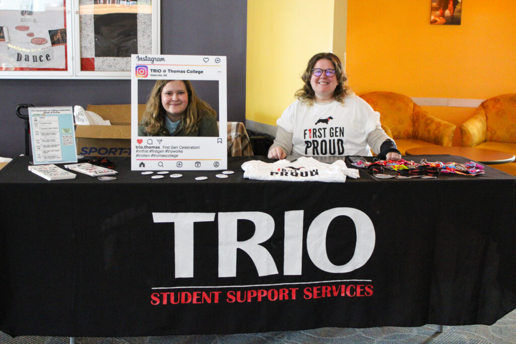 Two women sitting at a table with swag and a photo frame promoting the TRIO program at Thomas College