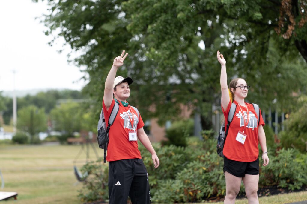 two students in red shirts waving to people arriving for move in and new terrier orientation