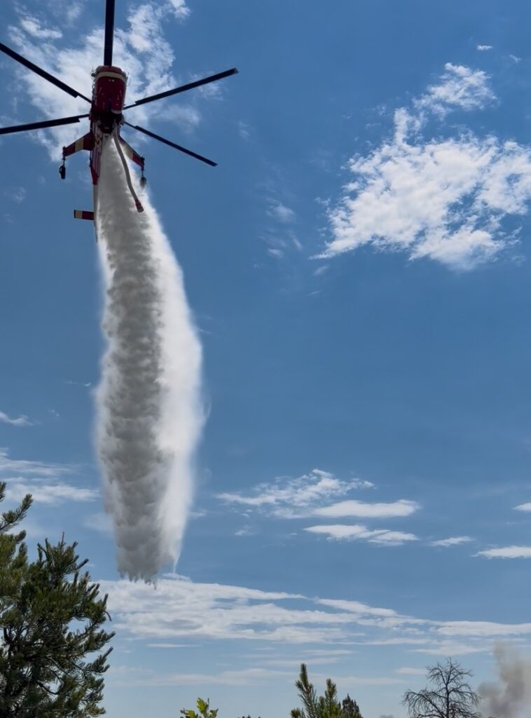 A view of a helicopter directly below it as water pours out of it to help fight a forest fire