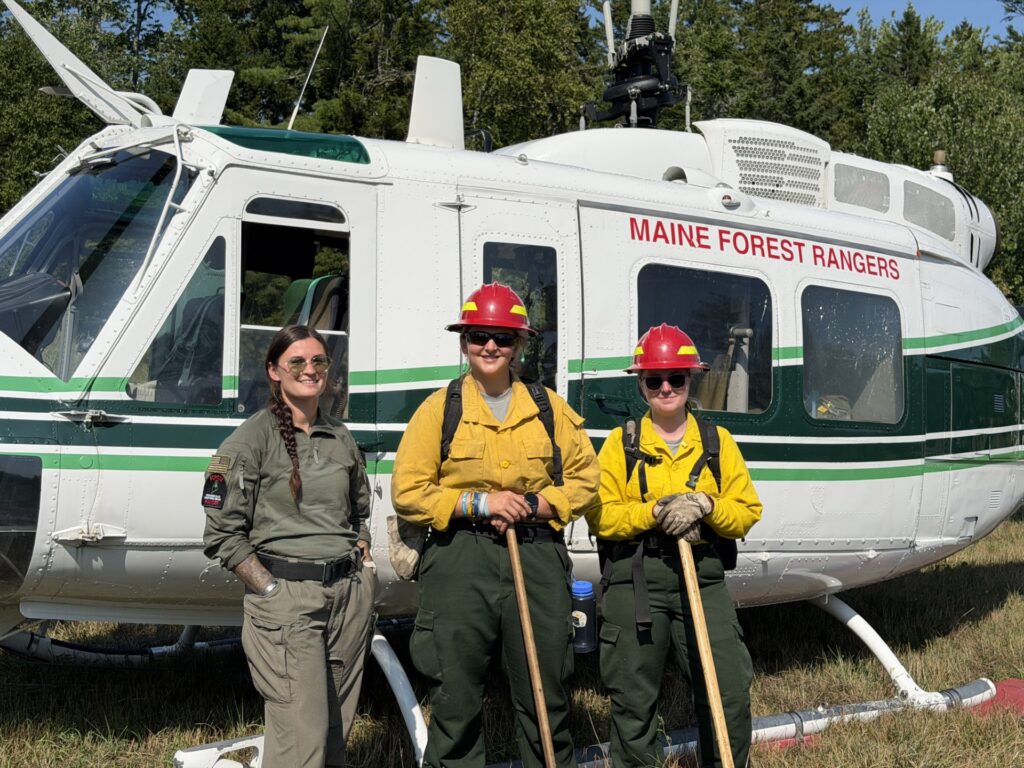 Three women dressed in fire fighting outfits stand in front of a Maine Forest Rangers helicopter